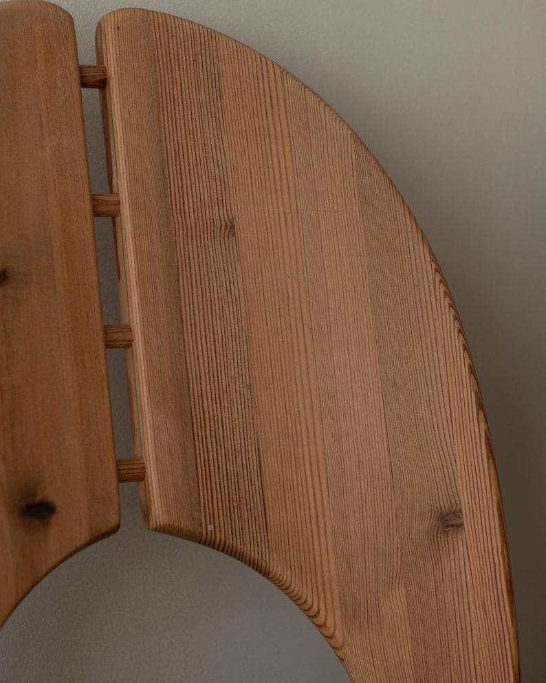 Puede incluir: Primer plano del respaldo de una silla de madera. El respaldo de la silla est&aacute; hecho de madera de color claro con vetas visibles. El dise&ntilde;o incluye una parte superior curva y listones verticales, mostrando una est&eacute;tica minimalista. La madera tiene nudos y variaciones naturales.