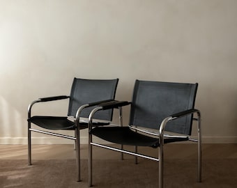 May include: Two black leather and chrome chairs. The chairs have a minimalist design with a rectangular back and seat, and curved armrests. The frames are made of shiny chrome, and the chairs are set against a neutral wall.