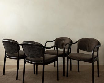 May include: A set of four dark brown chairs with curved black frames and rounded backrests. Each chair features a textured fabric seat and back panel, supported by four slender legs. The chairs are arranged in a group, showcasing a consistent design with integrated armrests that flow into the back structure.