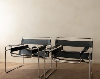 May include: Two lounge chairs featuring polished chrome tubular metal frames and black leather straps forming the seat, backrest, and armrests. The chairs have a low-profile design with continuous metal tubing that creates the base and frame.