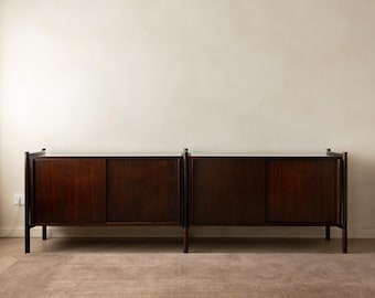 May include: A long, dark brown wooden sideboard featuring two distinct sections, each equipped with sliding doors. The piece is supported by slender, vertical legs at the corners and center, showcasing a smooth, polished finish and clean, linear construction.