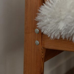 Puede incluir: Primer plano de un armaz&oacute;n de silla de madera con vetas visibles y fijaciones met&aacute;licas. Un coj&iacute;n de piel de oveja blanco y esponjoso descansa sobre el asiento. La silla tiene una est&eacute;tica natural y r&uacute;stica.