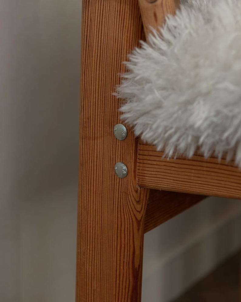 Puede incluir: Primer plano de un armaz&oacute;n de silla de madera con vetas visibles y fijaciones met&aacute;licas. Un coj&iacute;n de piel de oveja blanco y esponjoso descansa sobre el asiento. La silla tiene una est&eacute;tica natural y r&uacute;stica.