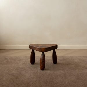Könnte beinhalten: Ein kleiner, dreibeiniger Hocker aus dunkelbraunem Holz. Die Sitzfläche hat eine abgerundete Dreiecksform mit glatten, geschwungenen Kanten. Er steht auf drei dicken, konisch zulaufenden zylindrischen Beinen, die eine stabile Basis bilden.