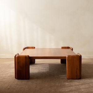 Op de afbeelding: Een vierkante salontafel gemaakt van middelbruin hout met een glad, plat oppervlak. De tafel heeft vier dikke, afgeronde hoekpoten die iets boven het tafelblad uitsteken, waardoor een duidelijk kader rond de randen ontstaat.