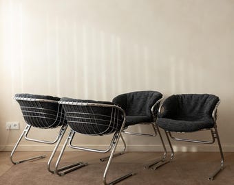 May include: Four chairs with polished chrome metal frames and dark gray upholstered seats. Each chair features a curved backrest constructed from a grid of metal wires, supported by a cantilevered base design with a sleek, metallic finish.