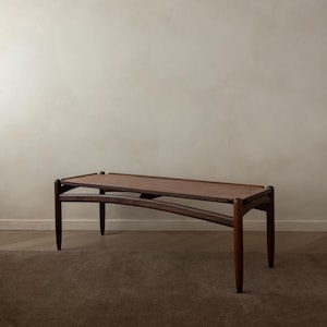 Puede incluir: Una mesa de centro de estilo moderno de mediados de siglo. La mesa tiene una tapa rectangular y un estante inferior, ambos de madera oscura. Las patas son anguladas y cónicas, y el diseño es minimalista y elegante. La mesa mide aproximadamente 122 cm de largo.