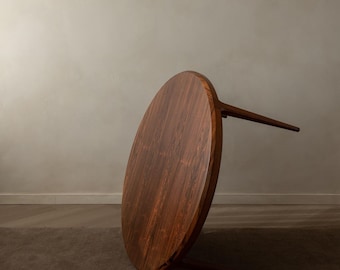 May include: A round, dark wood table with a rich, reddish-brown grain pattern. The table has a single leg extending from the center and two legs supporting the edge. The table is set on a brown carpet against a neutral wall.