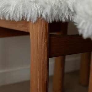 Puede incluir: Primer plano de una silla de madera con un asiento de piel de oveja blanca y esponjosa. Las patas de la silla son de madera marr&oacute;n claro con vetas visibles. El fondo es de color neutro.