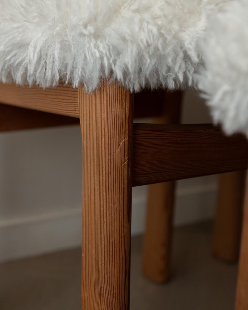 Puede incluir: Primer plano de una silla de madera con un asiento de piel de oveja blanca y esponjosa. Las patas de la silla son de madera marr&oacute;n claro con vetas visibles. El fondo es de color neutro.