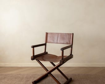 May include: A folding director chair constructed from dark brown wood with a smooth finish. The seat and backrest are made of a single piece of tan leather stretched across the frame. The chair features a cross-leg base and armrests.
