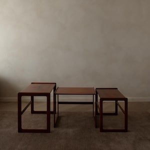 May include: Three wooden nesting tables in varying sizes. The tables are a rich, dark brown color with a simple, geometric design. The tables are arranged on a neutral-toned carpet, with a plain wall in the background.