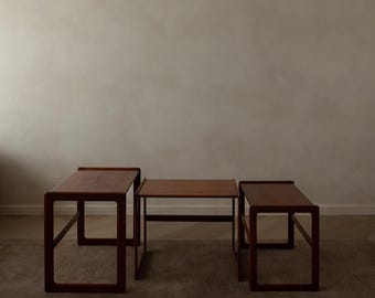 May include: Three wooden nesting tables in varying sizes. The tables are a rich, dark brown color with a simple, geometric design. The tables are arranged on a neutral-toned carpet, with a plain wall in the background.