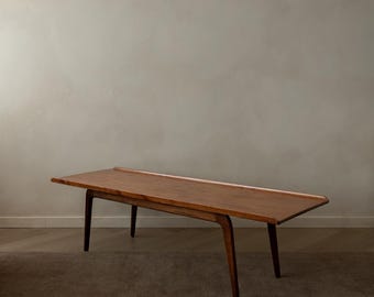 May include: A mid-century modern wooden coffee table with a rectangular top and four angled legs. The table is made of a warm-toned wood, likely walnut or teak, and has a simple, elegant design. The table measures approximately 60 inches long.