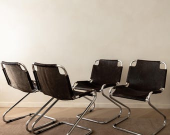 May include: A set of four chairs featuring continuous tubular chrome frames and dark brown cowhide leather seats and backrests. The chairs have a cantilevered design with a single curved metal base, providing a sleek and structural appearance.