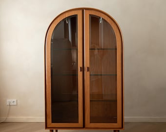 May include: A tall, arched wooden display cabinet featuring two glass-paneled doors with vertical wooden frames. The cabinet is supported by four slender, tapered wooden legs and contains internal glass shelving visible through the front.