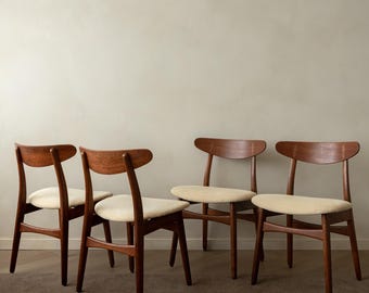 May include: Four mid-century modern chairs with curved backs and wooden frames. The seats are upholstered in a light beige fabric. The chairs are made of dark wood and have a minimalist design, suitable for a dining room or living space.