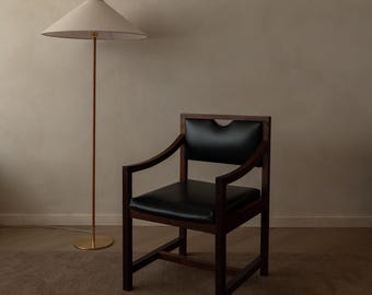 May include: A dark wood armchair with a black leather seat and backrest, featuring a curved top rail. Beside it stands a floor lamp with a thin metal pole, a circular brass base, and a wide, conical off-white fabric lampshade with a wood top.