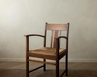 May include: A wooden armchair featuring a woven rush seat and a backrest with three vertical slats. The chair is constructed from dark brown wood and includes curved armrests, straight legs, and horizontal support stretchers between the legs.