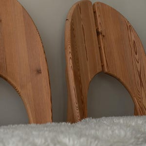Puede incluir: Dos cabeceros de madera de color marr&oacute;n claro con un borde superior curvo en forma de semic&iacute;rculo y un patr&oacute;n de veta de madera natural visible. Los cabeceros est&aacute;n colocados detr&aacute;s de una superficie blanca, texturizada y esponjosa.