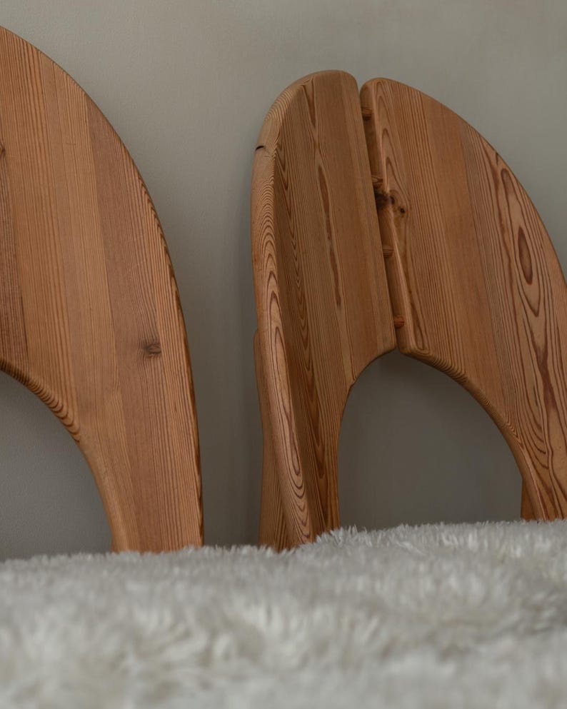 Puede incluir: Dos cabeceros de madera de color marr&oacute;n claro con un borde superior curvo en forma de semic&iacute;rculo y un patr&oacute;n de veta de madera natural visible. Los cabeceros est&aacute;n colocados detr&aacute;s de una superficie blanca, texturizada y esponjosa.