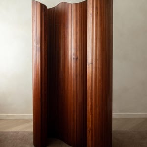 Puede incluir: Un biombo alto y autoportante construido con listones de madera verticales. La pieza presenta un acabado de grano de madera marrón cálido y rico, con una forma ondulada continua que le permite mantenerse en pie sobre sus bordes curvos.
