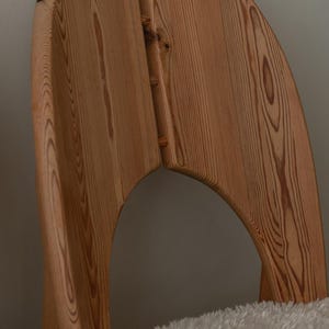 Puede incluir: Una silla de madera con respaldo curvo y acabado de vetas naturales. La silla tiene un dise&ntilde;o &uacute;nico con un recorte en la parte posterior y est&aacute; hecha de madera de color claro. La silla est&aacute; colocada sobre una superficie blanca y esponjosa.