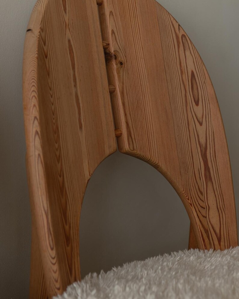 Puede incluir: Una silla de madera con respaldo curvo y acabado de vetas naturales. La silla tiene un dise&ntilde;o &uacute;nico con un recorte en la parte posterior y est&aacute; hecha de madera de color claro. La silla est&aacute; colocada sobre una superficie blanca y esponjosa.