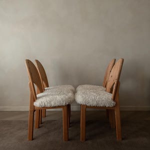 Puede incluir: Un conjunto de cuatro sillas de madera con marcos de color marr&oacute;n claro y respaldos curvos. Cada silla cuenta con un coj&iacute;n de asiento grueso de piel sint&eacute;tica blanca y peluda. Las sillas est&aacute;n dispuestas en dos pares frente a frente.