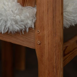 Puede incluir: Primer plano de una silla de madera con acabado marr&oacute;n claro. La silla tiene un coj&iacute;n blanco esponjoso. La veta de la madera es visible y la silla est&aacute; construida con uniones visibles. La silla parece ser un dise&ntilde;o moderno de mediados de siglo.