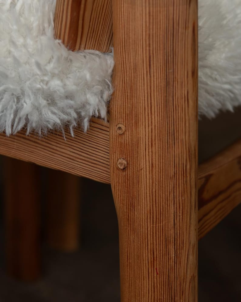 Puede incluir: Primer plano de una silla de madera con acabado marr&oacute;n claro. La silla tiene un coj&iacute;n blanco esponjoso. La veta de la madera es visible y la silla est&aacute; construida con uniones visibles. La silla parece ser un dise&ntilde;o moderno de mediados de siglo.