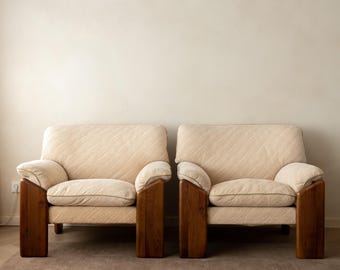 May include: Two matching armchairs featuring thick, dark brown wooden frames and light beige upholstered cushions with a subtle diagonal striped texture. Each chair has wide, block-style wooden armrests and a low-profile seat design.