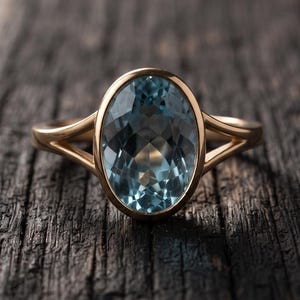 May include: A gold ring featuring a large, oval-cut, light blue gemstone set in a smooth bezel. The band has a split-shank design that tapers towards the centre setting, creating a clean, symmetrical look for this piece of fine jewellery.