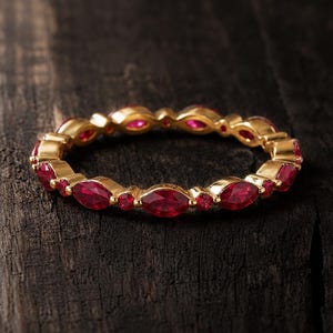 May include: A gold band ring featuring an alternating pattern of marquise-cut and round-cut red gemstones. The ring is constructed with a continuous metal setting that holds each stone securely in place, creating a circular design with a polished finish.
