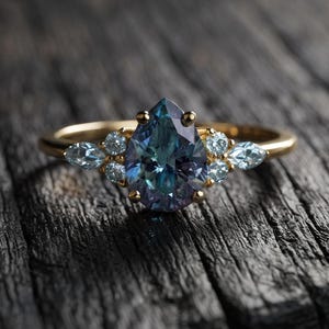 May include: A gold ring featuring a central pear-shaped, colour-changing gemstone that shifts between blue and purple hues. The centre stone is flanked by three small, clear, round-cut accent stones on each side, set in a polished gold band.