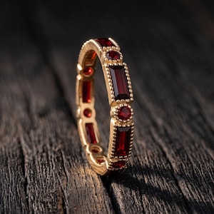 Puede incluir: Un anillo de oro con un patrón alterno de piedras preciosas rojas rectangulares de corte baguette y piedras rojas más pequeñas de corte redondo. El engaste de metal alrededor de cada piedra presenta una textura granulada detallada.