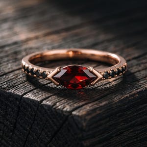 Peut inclure: Une bague en or rose ornée d'une pierre grenat rouge profond taille marquise au centre. L'anneau est agrémenté de petites pierres noires rondes serties de chaque côté de la tige, menant à la pierre centrale fixée par des griffes en métal poli.