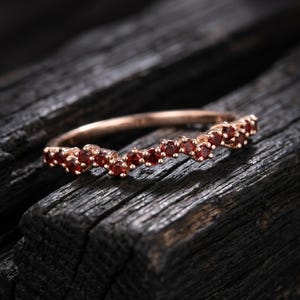 May include: A rose gold band ring featuring a chevron or V-shaped design set with a continuous row of small, round, deep red garnet gemstones. The metal has a polished finish, and the stones are secured in a prong setting along the band.