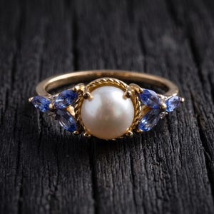 Puede incluir: Un anillo de oro con una perla blanca redonda central rodeada por un borde de metal trenzado. El engaste está decorado a ambos lados con tres piedras preciosas azules de talla marquesa dispuestas en forma de hoja sobre un aro fino.