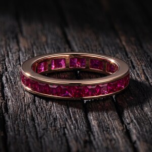Può includere: Un anello eternity in oro rosa con rubini taglio quadrato. La fascia dell'anello è di un colore oro caldo e lucido, e i rubini sono di un rosso intenso e vibrante. L'anello è esposto su una superficie di legno scuro e strutturato.