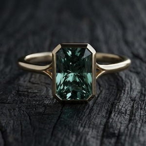 Puede incluir: Un anillo de oro con una piedra preciosa rectangular de color verde oscuro con talla esmeralda en un engaste de bisel. La banda es lisa y pulida, con un diseño de brazo dividido que se conecta al engaste central, mostrando una estructura limpia.
