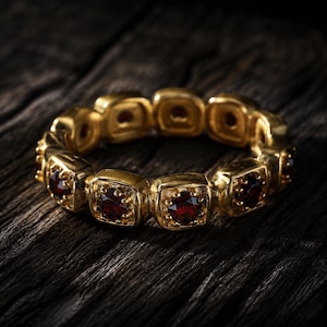 May include: A gold eternity ring featuring a series of square-shaped settings, each containing a round, deep red garnet gemstone. The band is constructed from connected metal links, with each setting displaying a faceted stone securely held in place.