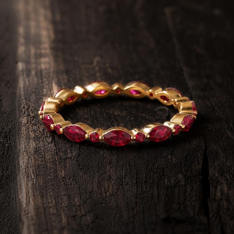 May include: A gold band ring featuring an alternating pattern of marquise-cut and round-cut red gemstones. The ring is constructed with a continuous metal setting that holds each stone securely in place, creating a circular design with a polished finish.
