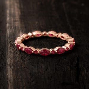 May include: A rose gold eternity band featuring an alternating pattern of marquise-cut and round-cut vibrant red rubies. The ring is crafted with a polished metal finish, showcasing a continuous design of gemstones set around the entire circumference.