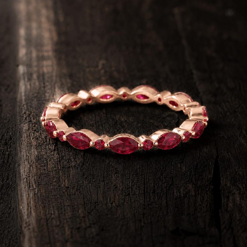 May include: A rose gold eternity band featuring an alternating pattern of marquise-cut and round-cut vibrant red rubies. The ring is crafted with a polished metal finish, showcasing a continuous design of gemstones set around the entire circumference.