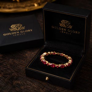 May include: A gold eternity ring featuring alternating marquise and round-cut red gemstones, presented inside a black jewelry box with the gold-printed text GOLDEN GLORY JEWELS and a diamond logo on the interior lid and the exterior box lid.