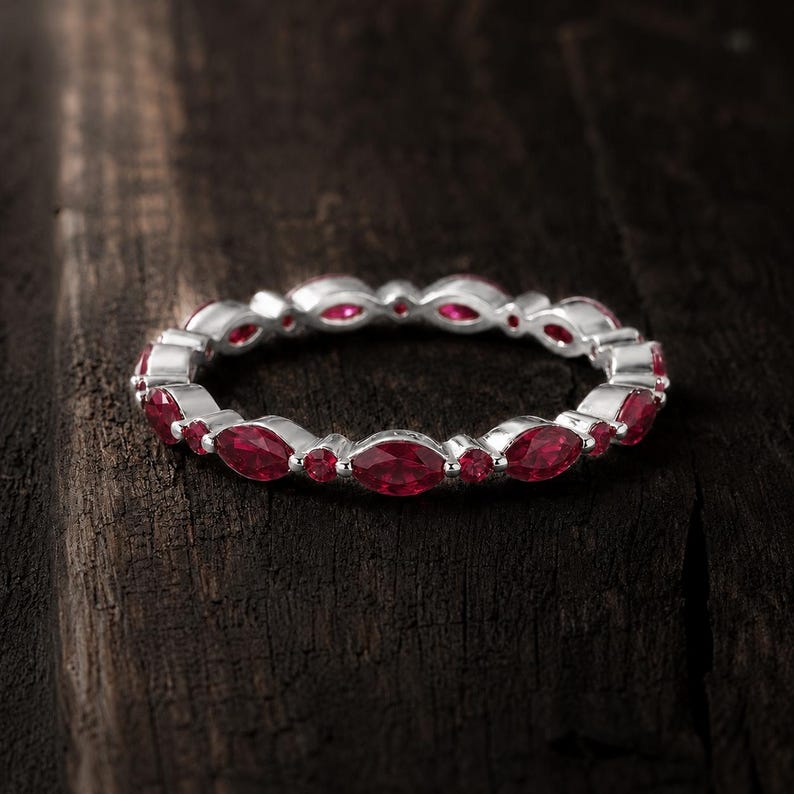 May include: A silver eternity ring featuring an alternating pattern of marquise-cut and round-cut red rubies set in a continuous band. The metal band has a polished finish, and the vibrant red gemstones are arranged symmetrically around the entire ring.