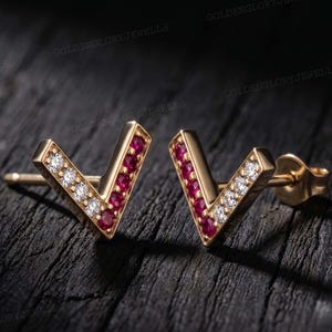 May include: A pair of V-shaped stud earrings crafted from gold-toned metal. Each earring features a row of round-cut clear white stones and a row of round-cut deep red stones set along the two arms of the V-shape, complete with post backings.