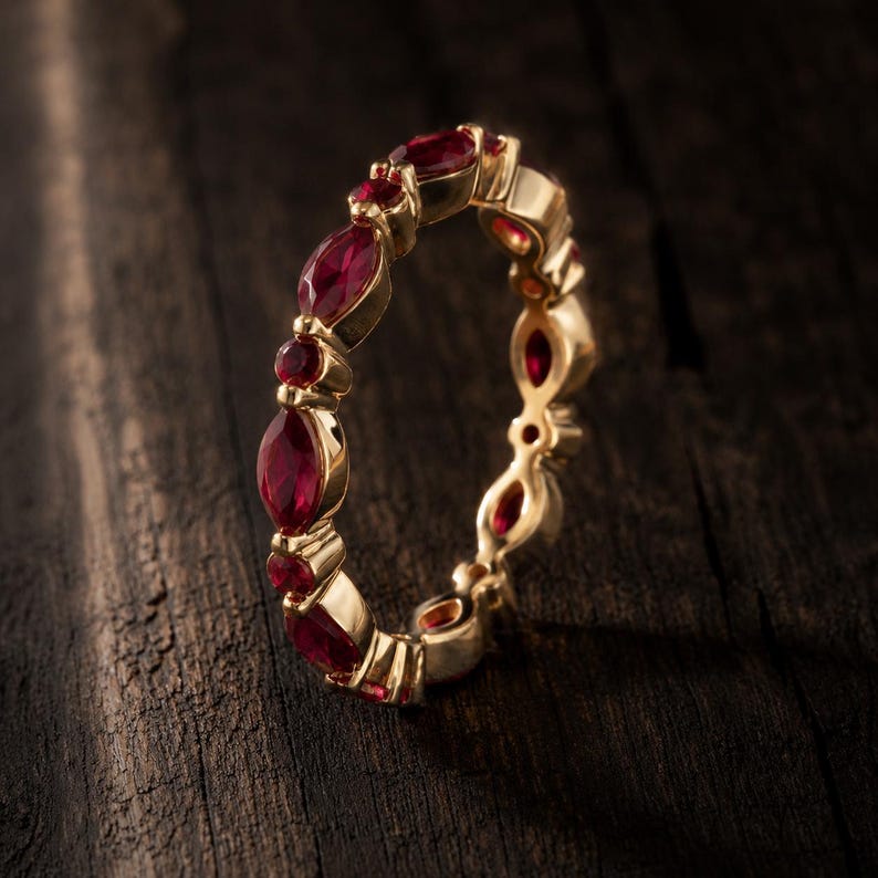 May include: A gold band ring featuring an alternating pattern of marquise-cut and round-cut red gemstones set in a continuous row. The metal has a polished finish, and the stones are secured by small prongs along the entire circumference of the ring.