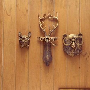 Puede incluir: Tres decoraciones de pared con cabezas de animales de color bronce: un gato con pajarita, un ciervo con pipa y un oso, todos con gafas. Las decoraciones están montadas en una pared de madera.